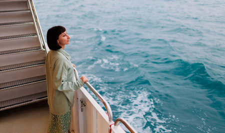 A young woman stands on the deck of a cruise ship and smiles. The girl smiles while traveling. A young beautiful brunette travels on a yacht. travel and adventure.の写真素材