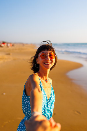 happy young woman pulls guy's hand. a beautiful girl in a dress walks along the beach, holding a guyの写真素材