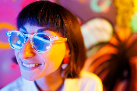 Portrait of a young girl in stylish glasses and fashionable clothes at a party. Beautiful sexy woman on a neon background, colored highlights on the girl's face with bright light.の写真素材