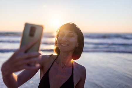 A beachgoer takes a selfie while enjoying the sunset by the ocean. The photo captures a joyful moment against the backdrop of waves and golden sunlight.の写真素材