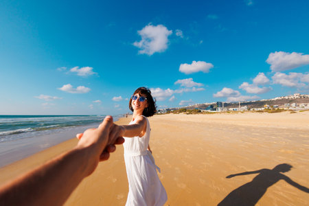 couple in love having fun on the beach. Follow me. happy young woman pulling guys hand. a beautiful girl in a dress walks along the beach, holding a guys hand, a romantic walk. Valentines Day.の写真素材