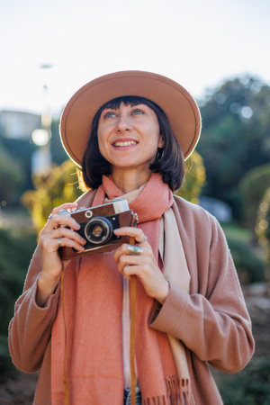 Fashionable look, young woman with a film camera wearing an elegant hat and coat. A hipster tourist woman holds a camera in her hands and takes a photo. film cameraの写真素材