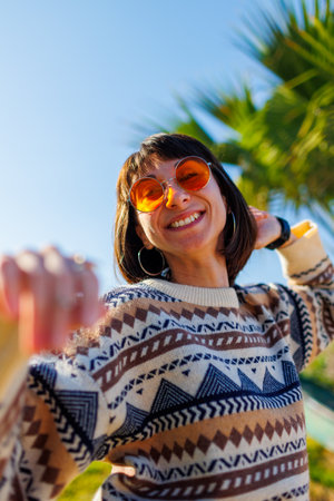 girl in a stylish knitted sweater and sunglasses looks at the camera and smiles. enjoys the holidays while standing on the street on a sunny day. real emotions.の写真素材