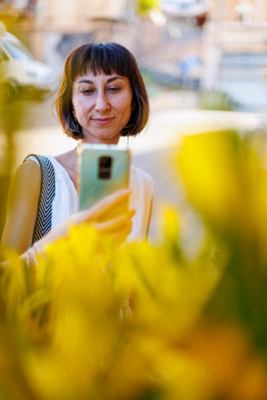young woman using a smartphone takes a photo of a decorative flower outdoors. A woman's hands are holding a mobile device, taking a photo of a beautiful plant on a sunny day. walk around the city.の写真素材