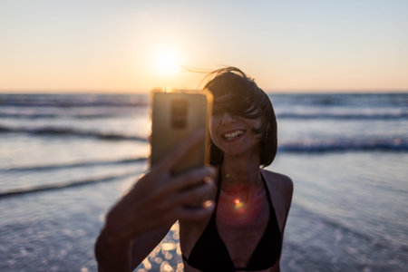 woman takes a selfie. A beautiful girl with a smartphone walks along the sea beach at sunset and...