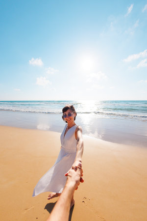 Follow me. happy young woman pulling friend's hand. couple in love having fun on the beach. a beautiful girl in a dress walks along the beach, holding her boyfriend's hand, a romantic walk.の写真素材