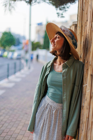 A young brunette smiles happily while standing in the city. A young stylish girl in a shirt, skirt and hat stands by the yard. Summer portraitの写真素材