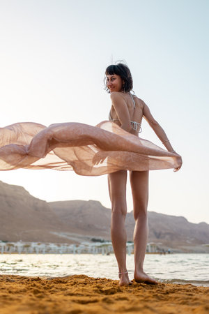 Beautiful girl on the beach enjoying the feeling of freedom at sunset. Woman in white swimsuit holding fluttering fabric playing in strong wind. Travel and vacation. Freedom concept.の写真素材