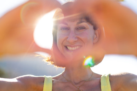 Smiling girl making love sign at sunset. romantic mood. Portrait of young smiling charming woman showing heart shape with hands, shining smile on face and looking through it.の写真素材