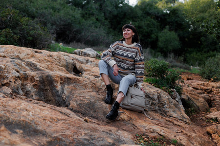 A smiling hiker in a sweater and with a backpack sits on a stone in the forest. female tourist sits on the ground and relaxes in nature in the mountains. hiking and walking in the forest.の写真素材