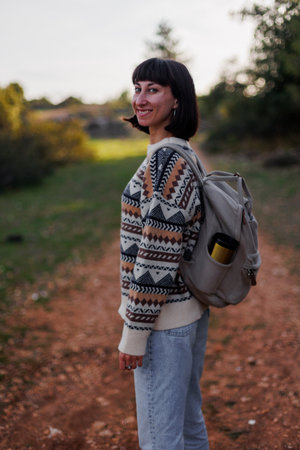 Smiling woman with a backpack hiking in nature. A beautiful female hiker stands on a mountain path and looks at the camera, smiling. Lifestyle, adventure, nature, active life.の写真素材