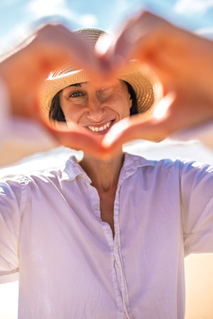 A cheerful young woman folds her hands in a heart shape against the backdrop of the ocean, enjoying the romantic atmosphere on the beach.の写真素材