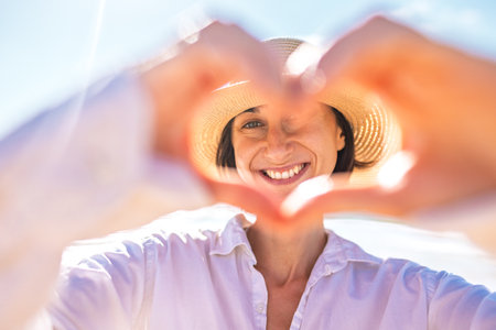 A cheerful young woman folds her hands in a heart shape against the backdrop of the ocean, enjoying the romantic atmosphere on the beach. She's in a romantic mood.の写真素材