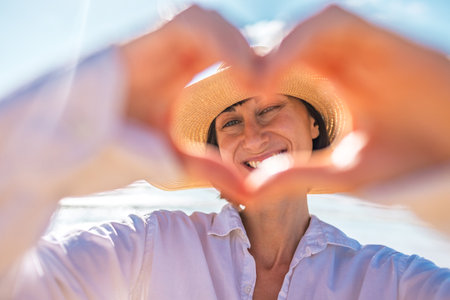 A girl looks into the camera and makes a beautiful heart with her fingers. A cheerful girl with a romantic mood against the backdrop of the sea and sky. Romantic mood.の写真素材