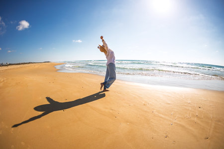 A happy, beautiful young woman strolls along the beach. A carefree girl strolls along the beach on a sunny day. A slender girl in a white shirt on the beach.の写真素材