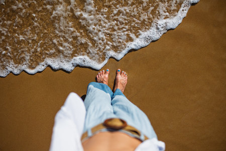 A girl stands barefoot on the sand, watching the waves touch her feet. POV shot, close-up. The joy of walking barefoot in the seawater. A moment of happiness. Bare feet in the water.の写真素材