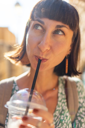 Portrait of a happy young woman drinking a cocktail through a straw while standing on a city street on a sunny summer day. A cheerful lady enjoys a cold drink on a city street. Takeaway coffee.の写真素材