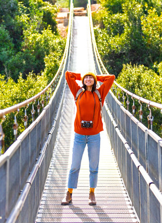 young woman with a camera and a backpack enjoys a walk along the suspension bridge. Happy female...