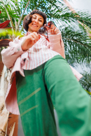 young and beautiful smiling girl in a fashionable pink shirt and sunglasses in her hands. Carefree woman posing outdoors on a sunny day near palm trees. Positive model outdoors. Cheerful and happy.の写真素材