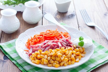 Salad with fresh carrots, sausage and corn on a white plate on a wooden table, horizontalの写真素材
