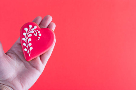 Gingerbread in the form of a heart with a beautiful pattern in female hands on a red background,...