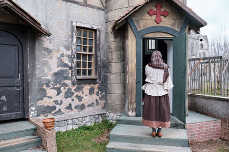 Young woman parishioner stands with her back and prays at the door of a medieval church. Halloween gothic vintage costumeの写真素材