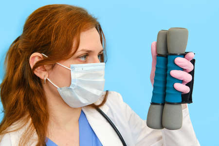 Woman doctor holds dumbbells for sports on a blue background, close-up.の写真素材