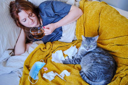 Cat in bed of a sick woman drinking tea. Woman with a coronavirus drinks tea while lying on a bed next to a catの写真素材