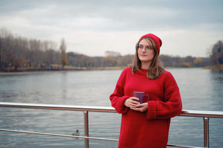 Adult woman by the autumn river with a cup of coffee in her hands, lifestyleの写真素材