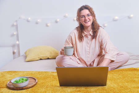Happy woman studying online with laptop on home bed. Student remotely learns during isolation due to coronavirusの写真素材