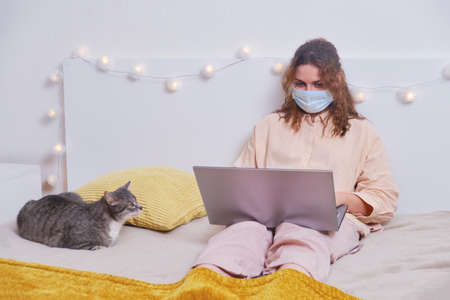 Young woman works in a face mask from home. A girl in quarantine due to coronavirus with a laptop and a pet, home lifestyle.の写真素材