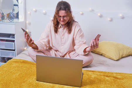 Problems of remote work via the Internet during isolation due to quarantine. Unhappy woman crying with electronic devices in her hands and a laptop on the home bed.の写真素材
