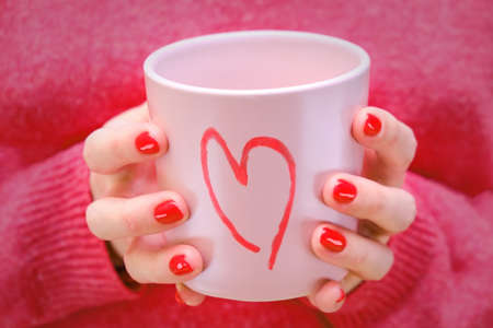 A woman holds a pink cup with a painted heart. The girl in the red sweater with a romantic mug.の写真素材
