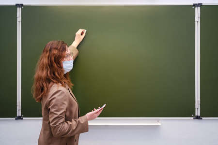 A teacher in a medical mask with a phone in hand writes a text on a school blackboard, copy space. School quarantine education conceptの写真素材