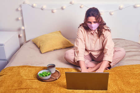 Redhead woman in medical mask works on laptop in yellow bed. Girl works at the computer online during isolation due flu virus, conceptの写真素材