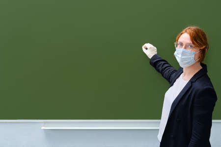 A teacher in a medical mask writes with chalk on an empty blackboard, copy space. A woman in a suit stands in a school classroom, close-upの写真素材