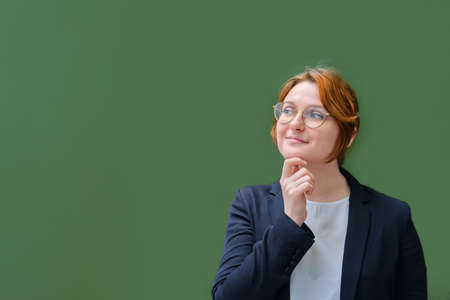The teacher stands next to an empty school blackboard, copy space for text. Happy woman teacher smiles next to an empty blackboardの写真素材