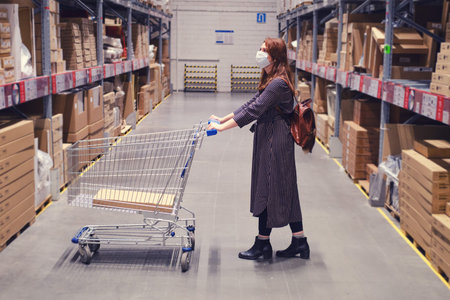 A young woman in a medical mask with a cart in a store warehouse