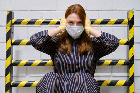 A woman in a medical glove holds her head against the background of a fire barrier with yellow-black stripes. Quarantine and isolation from viruses and diseases, conceptの写真素材