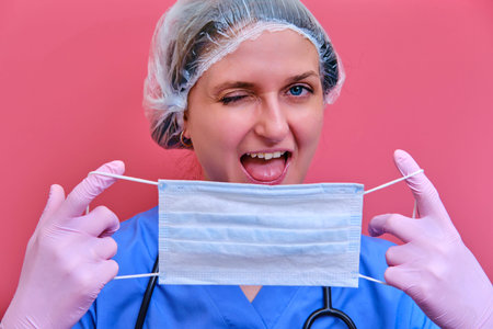 A happy female doctor holds a medical mask and winks, copy space for the inscription. Woman nurse with a protective mask in her hands on a pink background.の写真素材