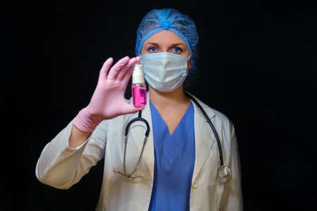 Doctor in a medical mask with hand sanitizer, copy space. A nurse holds a disinfectant in her hand on a black background. Concept of medical work in the coronavirus epidemicの写真素材