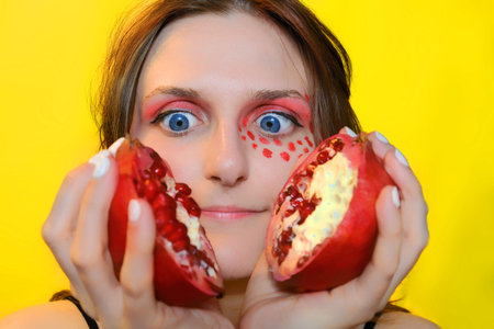 A young woman with two halves of red fruit. Female face and red garnet close-up. Girl and fruit on a...