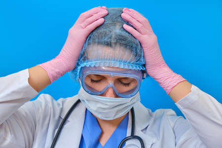 The doctor holds his hands over his head on a blue background, headache. Nurse in medical uniform - stress and fatigue due coronavirusの写真素材