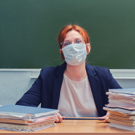Woman teacher sitting in a medical mask at a school desk, close up. Sad school teacher because of...