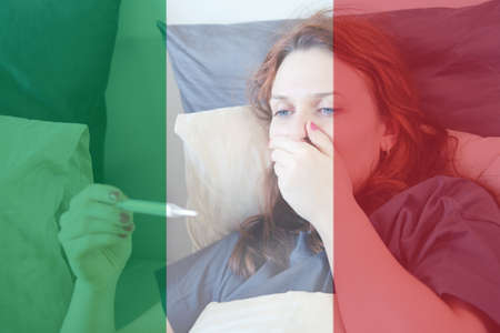 A woman with a thermometer lies sick with coronavirus in bed against the background of the flag of Italyの写真素材