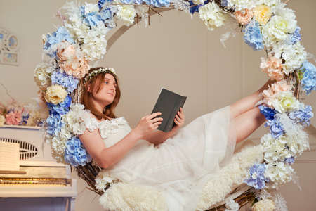 A young woman lies with a book in flowers. Studio portrait of happy woman reading book in white dressの写真素材