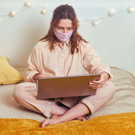 Woman in face mask works online on the yellow bed. Young girl in a medical mask with a computer at a remote workの写真素材
