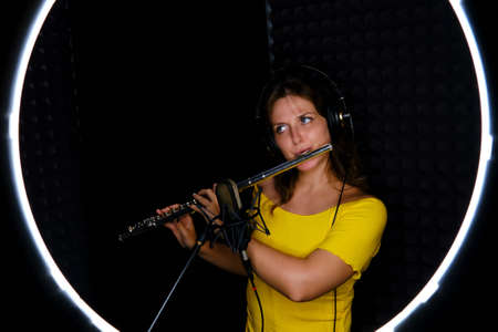 Woman flutist in headphones playing the large orchestral flute. Female flute player plays in recording studio, copy space. Record wind musical instruments with a professional microphone, ring light.の写真素材