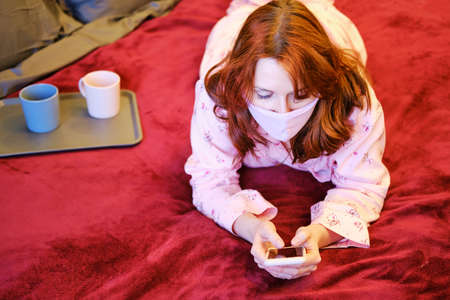 A woman lies on a bed with a phone during a coronavirus disease, face closeup. Girl in pink pajamas and a protective mask during an illness with the covid-19 flu virus, portraitの写真素材