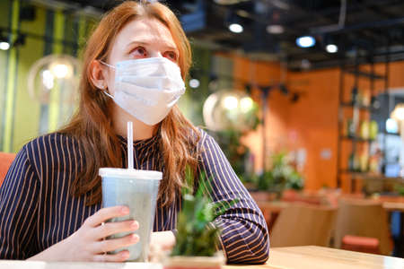 A woman in a medical mask sits at a table in a cafe. Girl with a coronavirus flu in a restaurantの写真素材